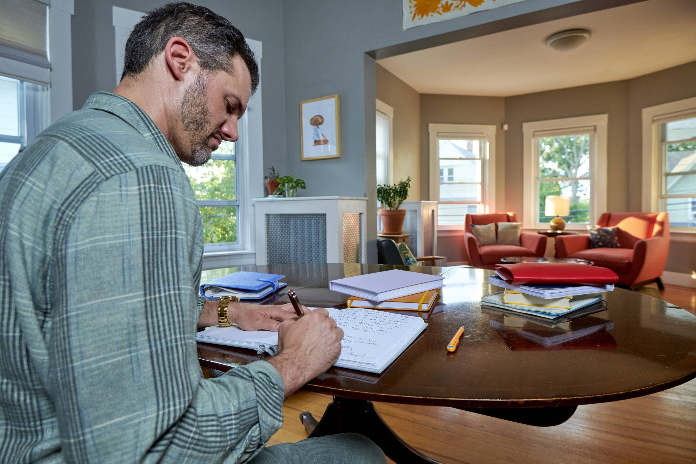 Marcus Berley writing in a notebook at a dark wood table, in a sunlit room with two red armchairs